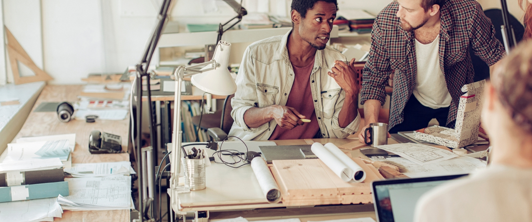 Two people sit at a drafting desk and talk to each other about a project. Rolled up drafting plans sit on the desk, along with a white desk lamp.
