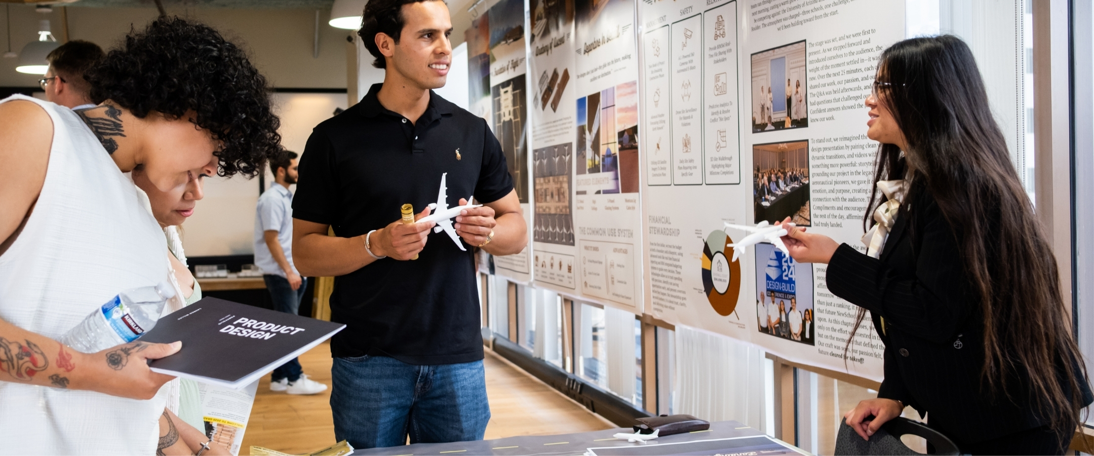 Two students talk to each other about a project while one holds a small prototype of an airplane. Another student stands to the left holding a brochure that says product design