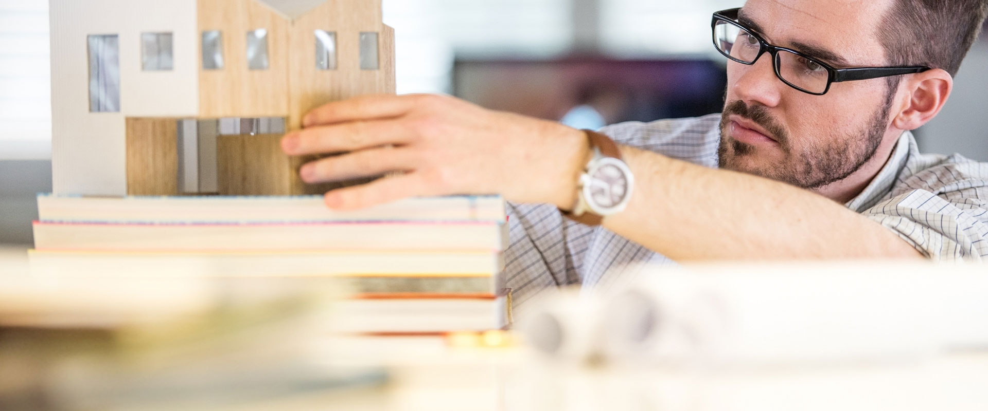 A man wearing glasses balances a wooden building model on a stack of books