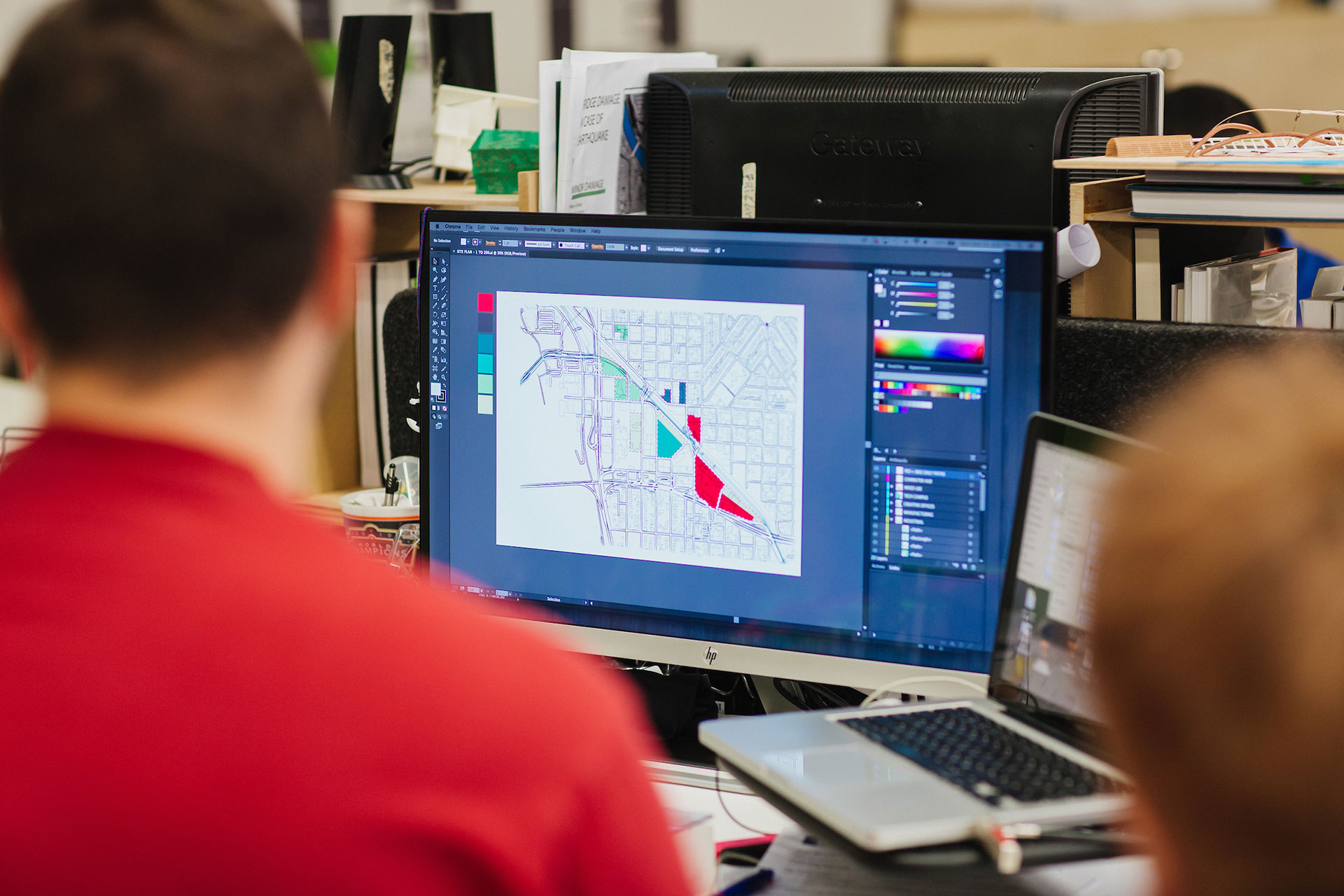 Two people view a computer screen displaying a digital map being edited in design software, with color panels and tools visible around the workspace.