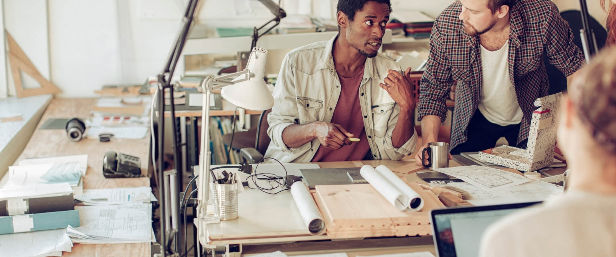 B.-Architecture-Careers-Photo (1) Two people sit at a drafting desk and talk to each other about a project. Rolled up drafting plans sit on the desk, along with a white desk lamp.